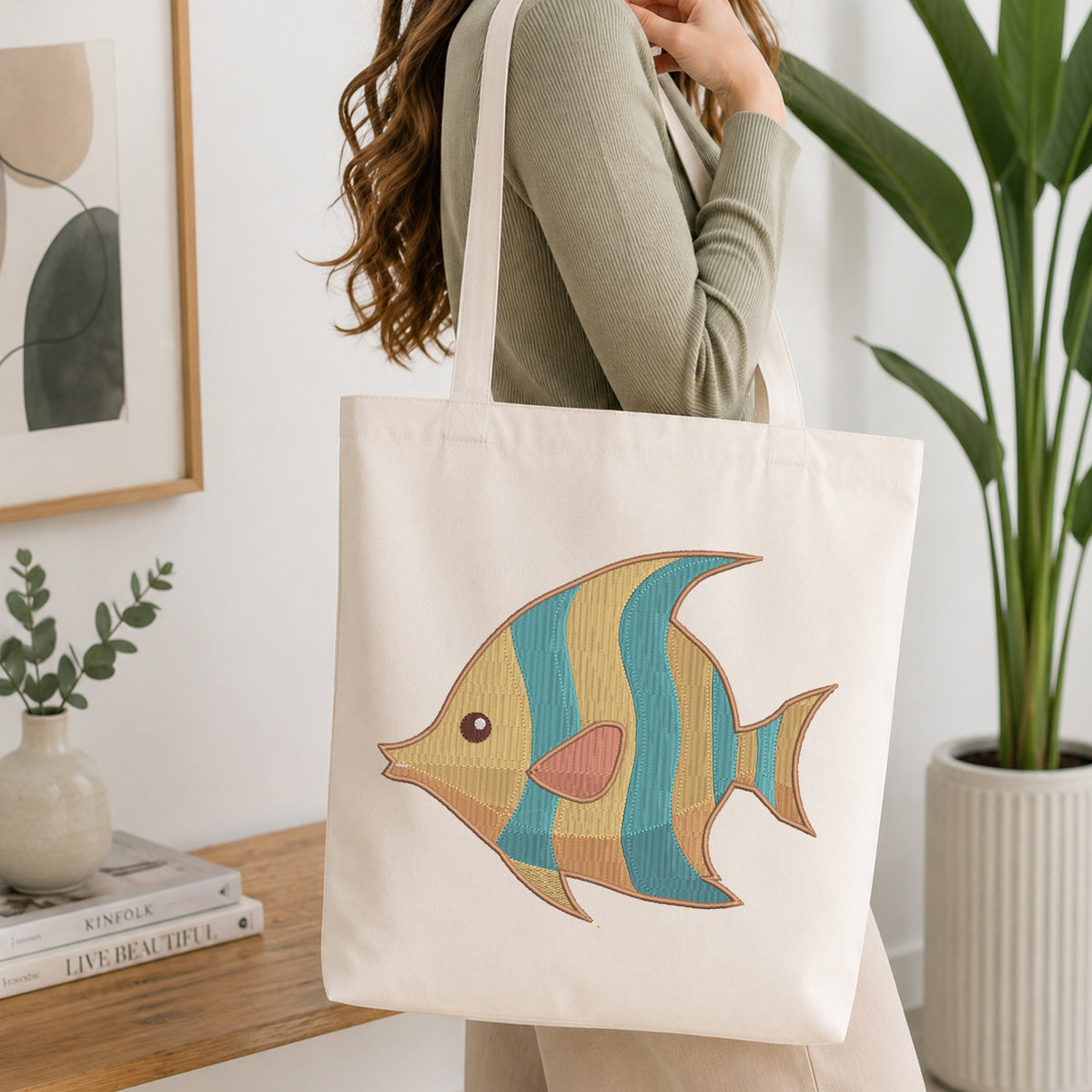 A woman wearing a sage green ribbed sweater carries a cream canvas tote bag with a colorful embroidered tropical fish design. The background is a bright, modern room with a wooden table, books, a framed print, and a potted plant.