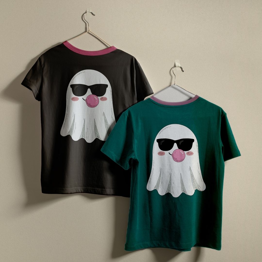 Two children's ringer t-shirts hanging on hangers against a plain beige wall. One shirt is black with a pink collar and features a white ghost graphic wearing black sunglasses and blowing a pink bubble.