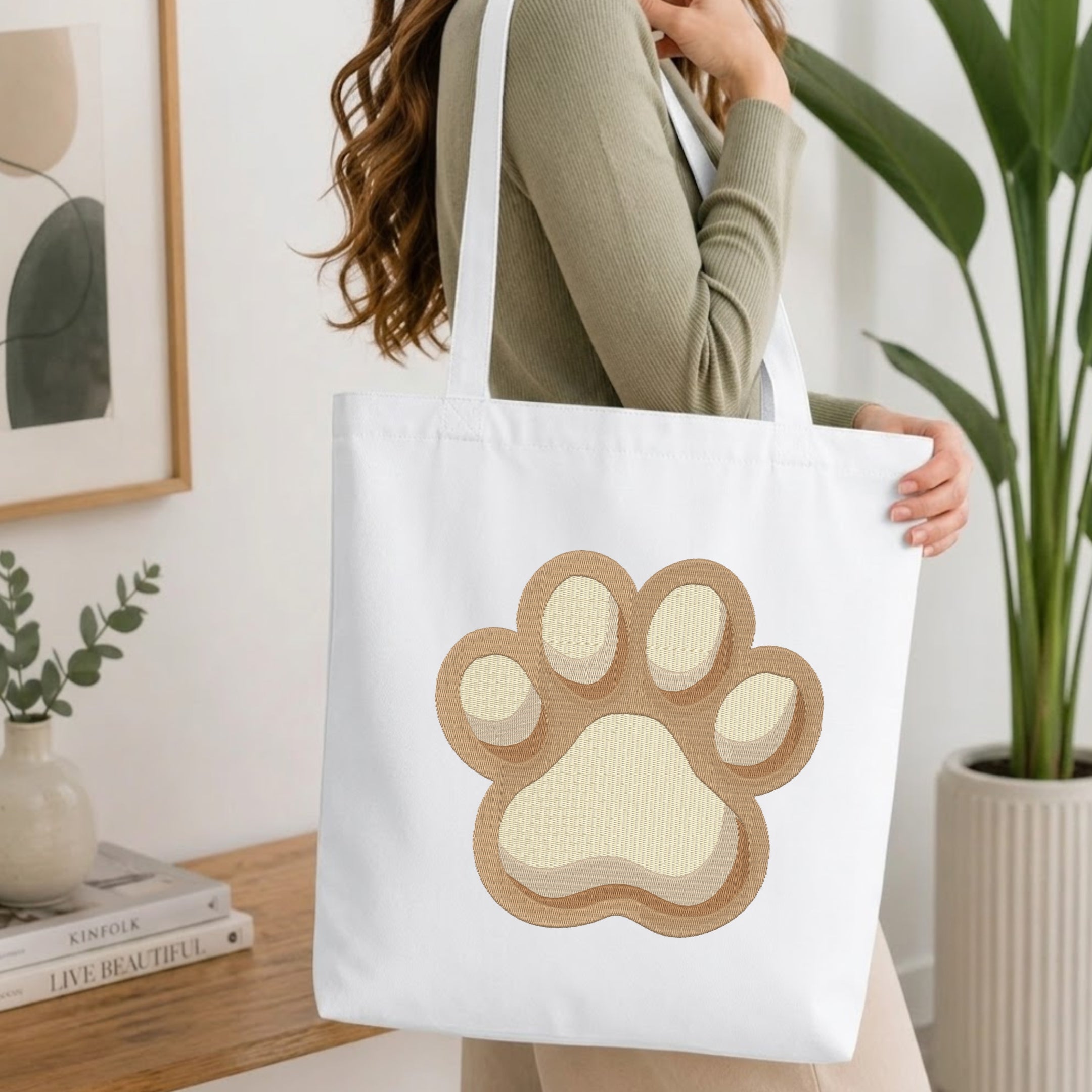 A woman wearing a sage green ribbed long-sleeve top carries a white canvas tote bag with a large applique-style paw print on the front. The paw print features a tan outer border and a cream center.
