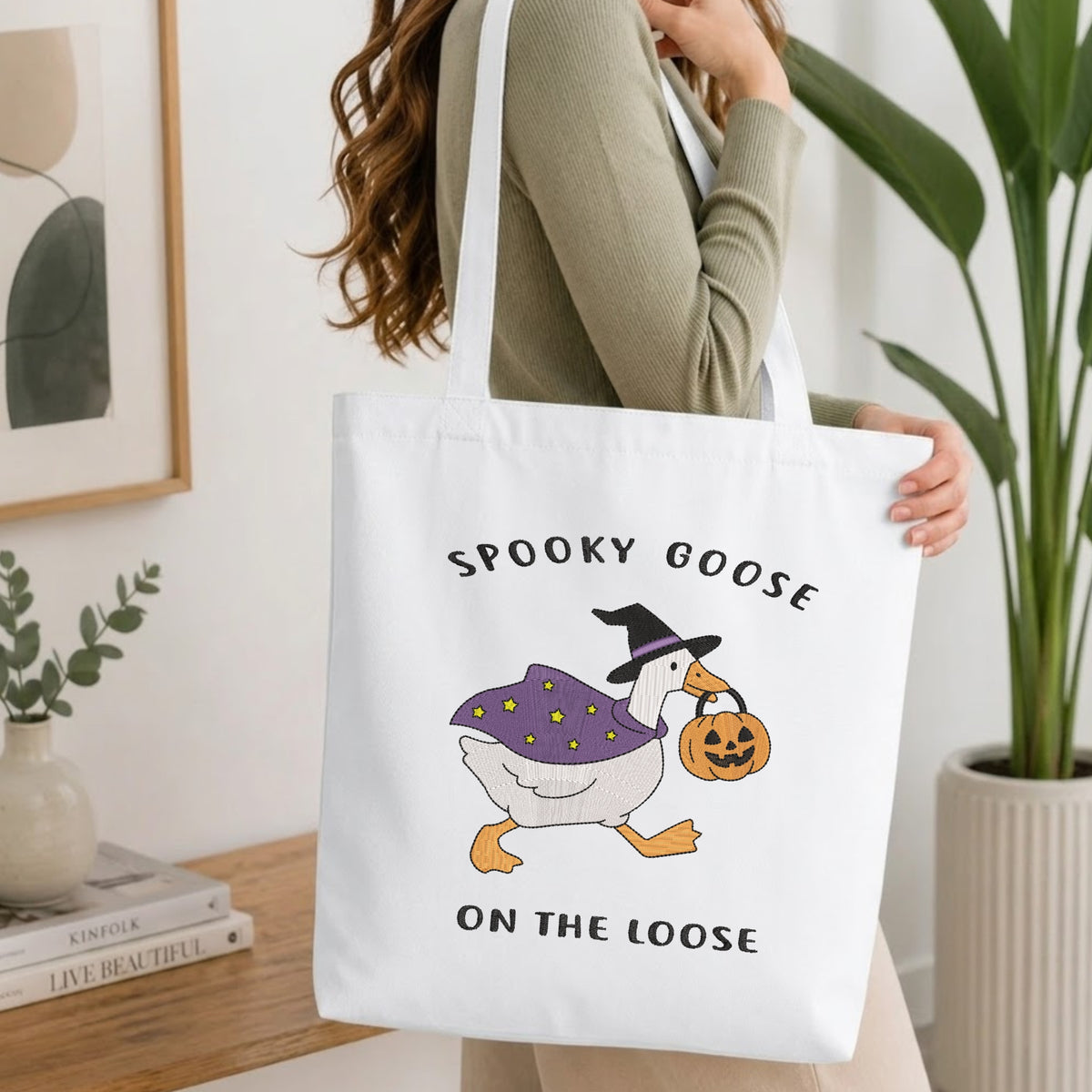 Woman carrying a white natural canvas tote bag featuring an embroidered "Spooky Goose" design in a bright, modern interior setting