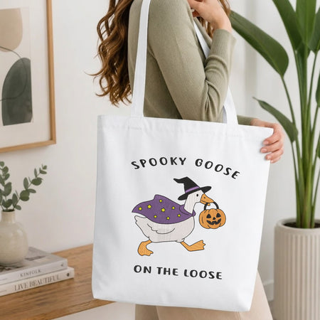 Woman carrying a white natural canvas tote bag featuring an embroidered "Spooky Goose" design in a bright, modern interior setting