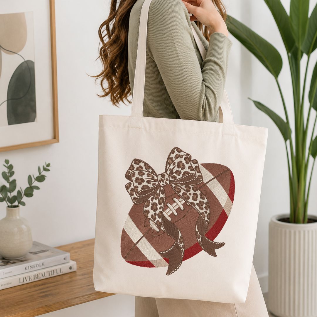 A woman wearing a sage green ribbed sweater carries a cream canvas tote bag with a football-themed bow and heart graphic. The background shows a cozy indoor setting with a wooden table, books, a framed print, and a potted plant.