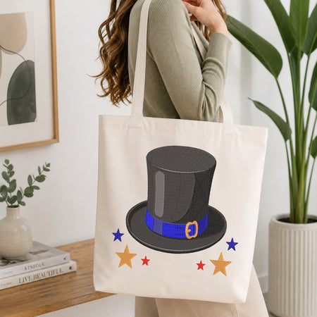 A woman wearing a sage green ribbed sweater carries a cream canvas tote bag with a printed top hat and stars. The tote bag features a black top hat with a blue band and gold buckle, surrounded by red and blue stars.