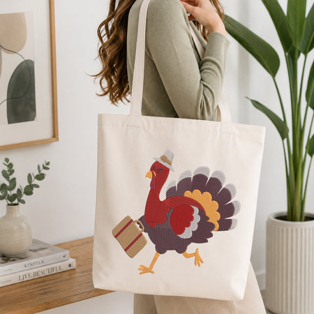 A woman wearing a sage green ribbed sweater carries a cream canvas tote bag with a colorful cartoon turkey graphic. The scene is set in a bright, modern room with a wooden table, books, a small vase, and a potted plant.