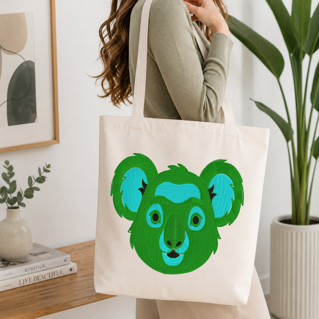 A woman wearing a ribbed sage green top carries a cream canvas tote bag featuring a stylized green and blue koala face graphic, set in a bright, modern room with a wooden table, books, and a potted plant.