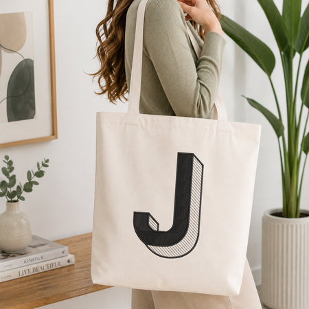 A woman wearing a sage green ribbed long-sleeve top carries a cream canvas tote bag with a large black stylized letter 'J' printed on the front, set in a bright, modern room with a wooden table, framed art, and a potted plant.