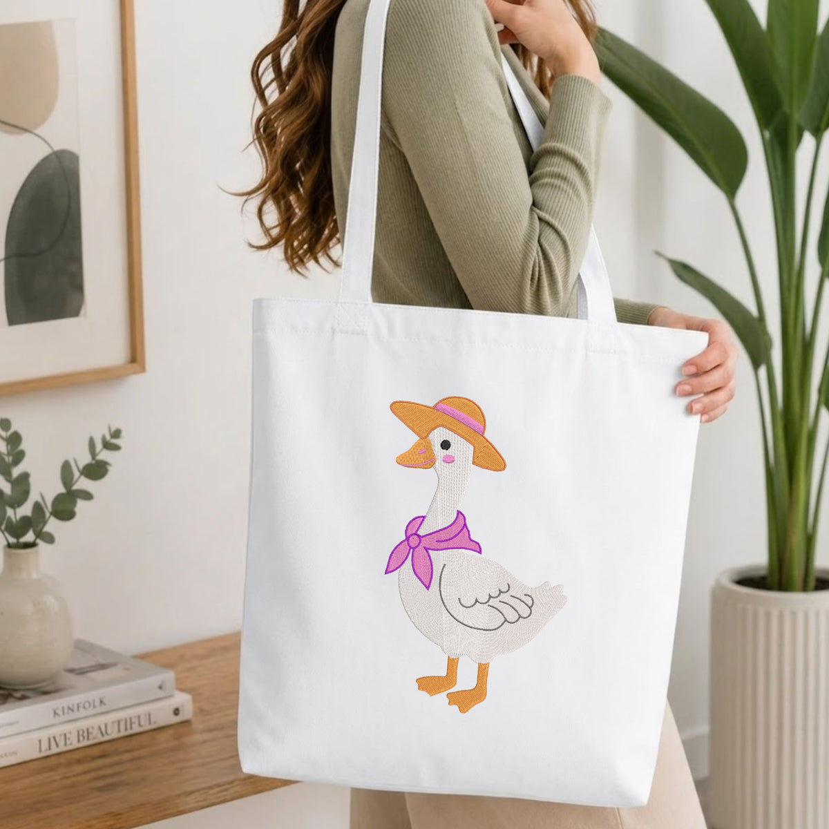 Woman carrying a white natural canvas tote bag featuring an embroidered cute duck in a straw hat design in a bright, modern interior setting