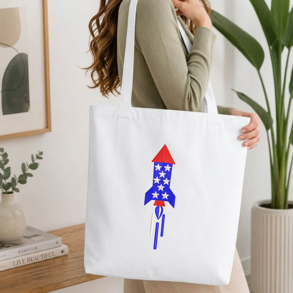 Woman carrying a white natural canvas tote bag featuring an embroidered patriotic space rocket star pattern design in a bright, modern interior setting.