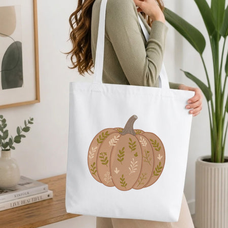 Woman carrying a white natural canvas tote bag featuring an embroidered leaf-patterned pumpkin design in a bright, modern interior setting.   