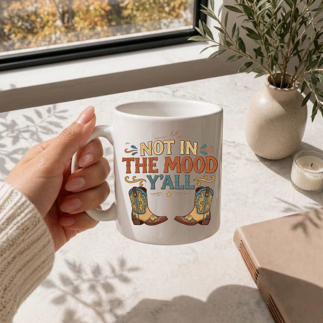 Hand holding a mug with 'Not in the Mood Y'all' text and cowboy boots design on a table with a window and plant in the background.