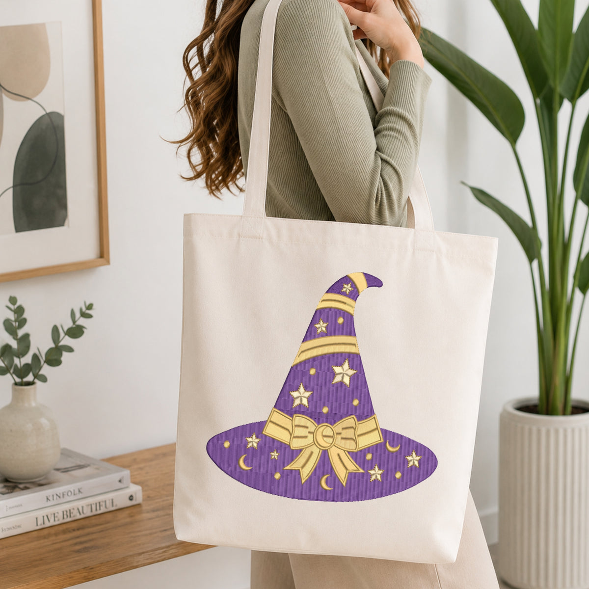 Flat-lay mockup showing a folded white t-shirt featuring a purple witch hat embroidery design with gold star and bow accents, set against a cozy white knitted blanket background.