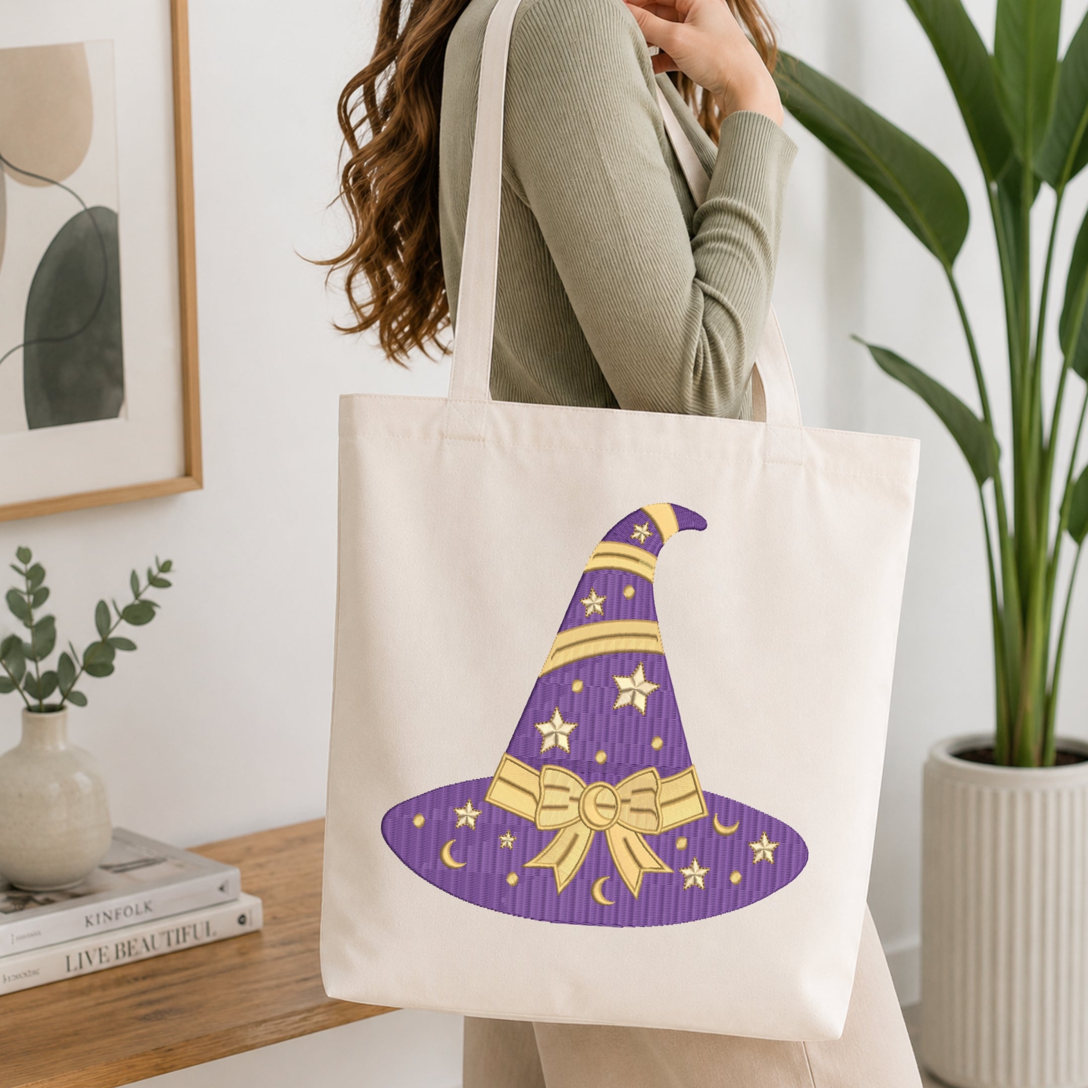 Flat-lay mockup showing a folded white t-shirt featuring a purple witch hat embroidery design with gold star and bow accents, set against a cozy white knitted blanket background.