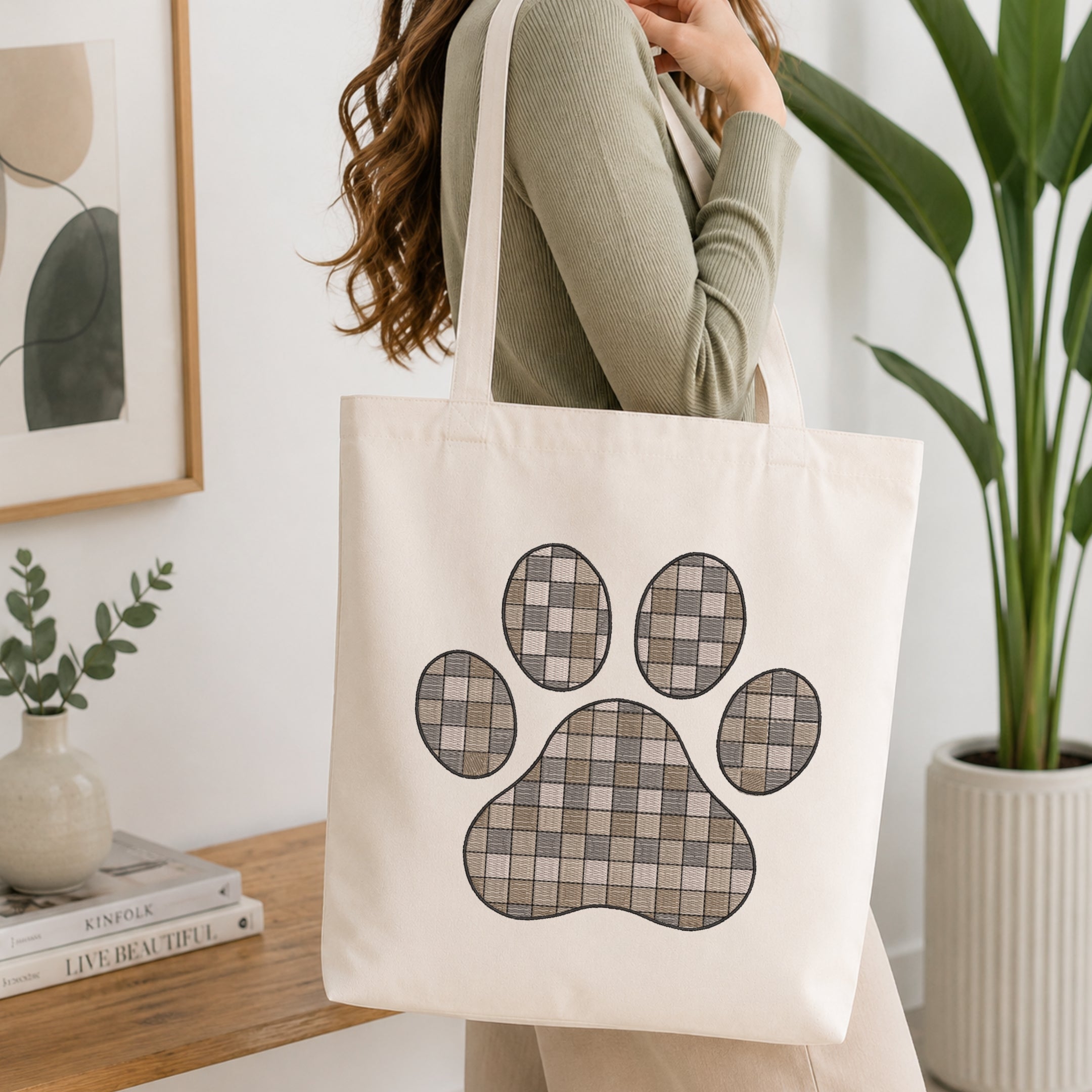 A woman wearing a sage green ribbed top carries a cream canvas tote bag with a large paw print graphic in a gray and beige plaid pattern. The scene is set in a bright, modern room with a wooden table, books, a small plant, and a potted green plant in the background.