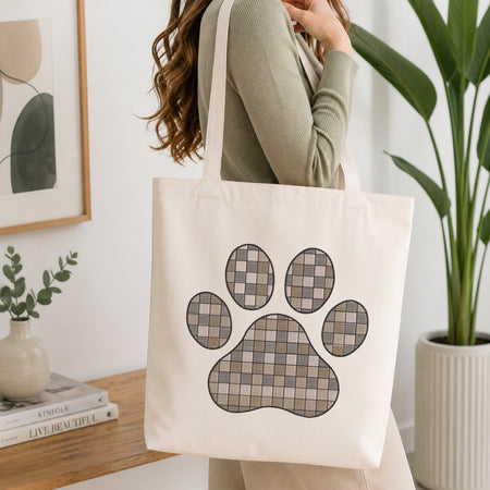 A woman wearing a sage green ribbed top carries a cream canvas tote bag with a large paw print graphic in a gray and beige plaid pattern. The scene is set in a bright, modern room with a wooden table, books, a small plant, and a potted green plant in the background.
