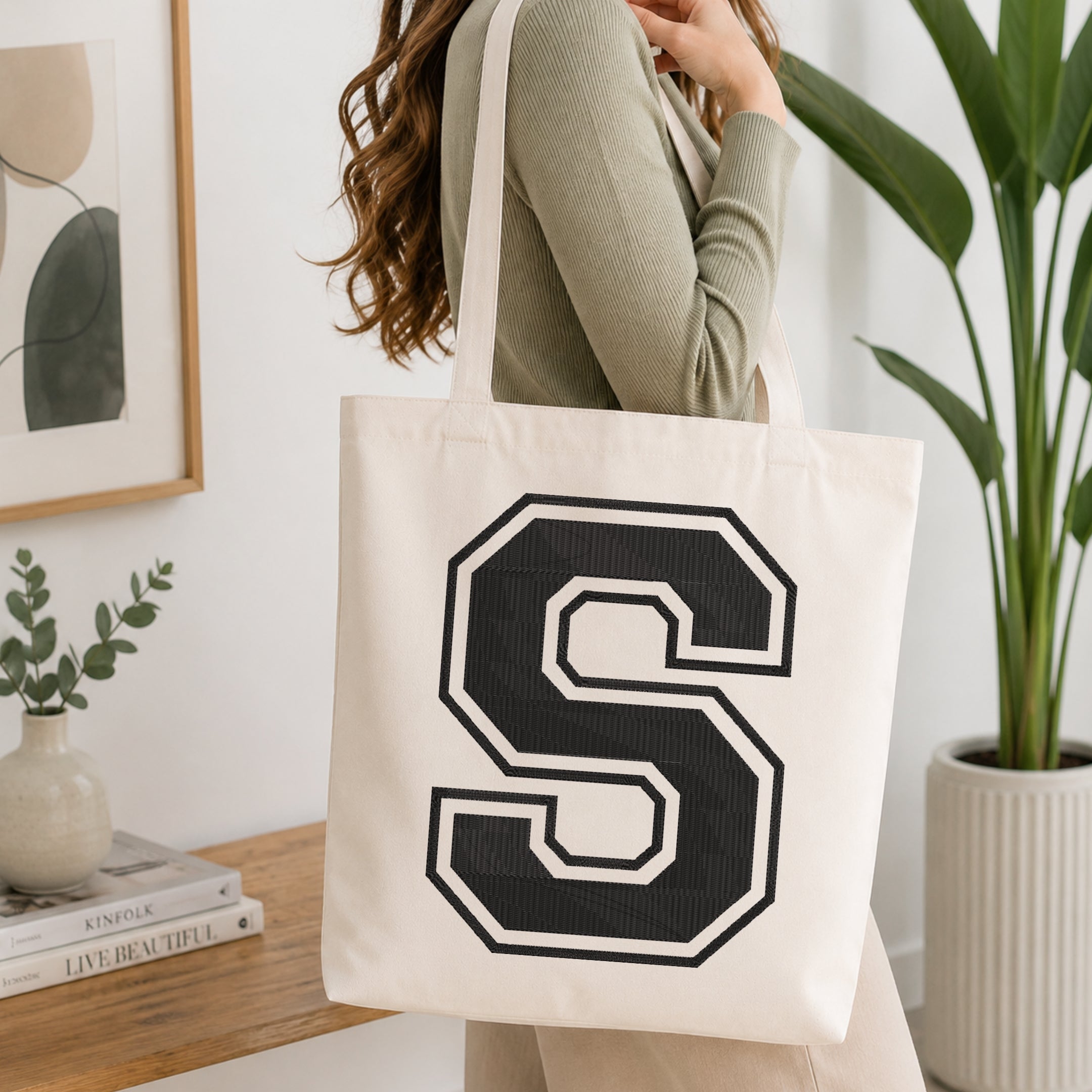 A person carrying a white canvas tote bag featuring a large embroidered varsity block letter 'S' in black with a white outline, set in a bright room with green house plants.
