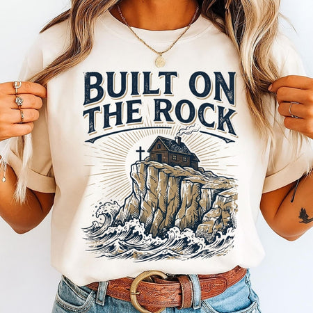 Person wearing a t-shirt with 'Built on the Rock' design and illustration of a house on a cliff.