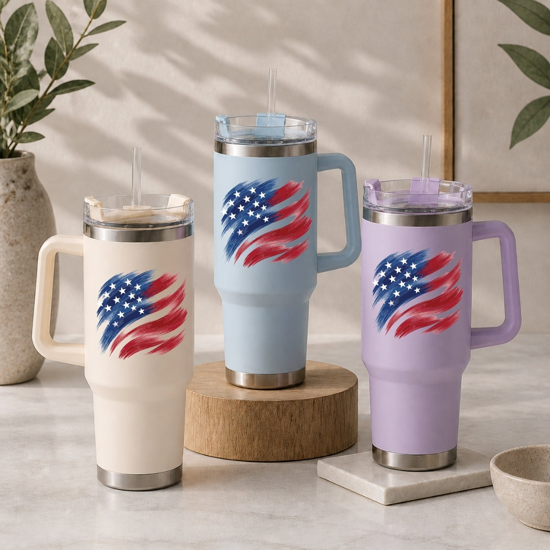 Three stainless steel tumblers in cream, light blue, and light purple, each decorated with a stylized brushstroke American flag graphic.