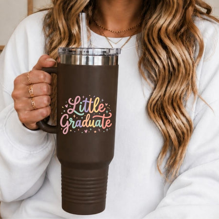 A person in a white sweatshirt holds a dark brown insulated tumbler with a clear lid and straw. The tumbler has the words "Little Graduate" in pastel script with small colorful stars and hearts.