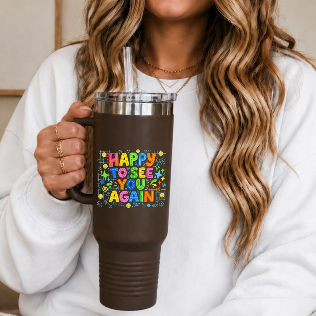 A person in a white sweatshirt holds a dark brown insulated tumbler with a clear lid and straw. The tumbler has a colorful retro-style graphic that reads 'HAPPY TO SEE YOU AGAIN' with small yellow stars and dots.
