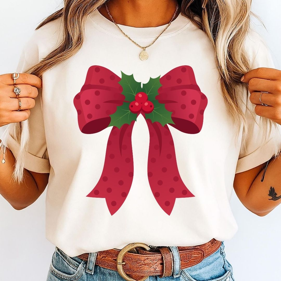 A woman wearing a cream-colored t-shirt with a large red bow and green holly graphic on the front, paired with blue jeans and a brown belt.
