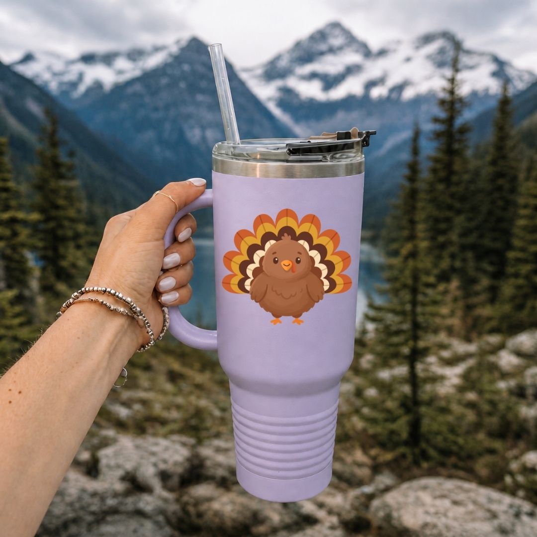 A hand holds a lavender insulated tumbler with a clear straw in front of a mountain and forest background. The tumbler has a cartoon turkey graphic on the front.