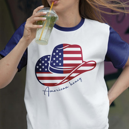 Person wearing a white t-shirt with an American flag design and 'American honey' text, holding a drink.