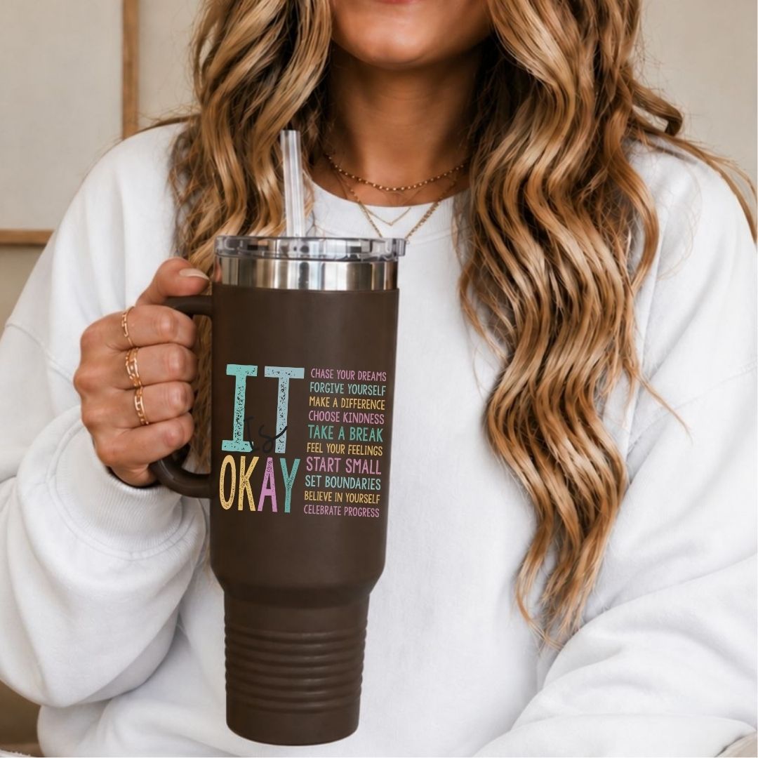 A woman in a white sweatshirt holds a dark brown insulated tumbler with a clear lid and straw. The tumbler has colorful text that reads 'IT OKAY' with smaller motivational text underneath.