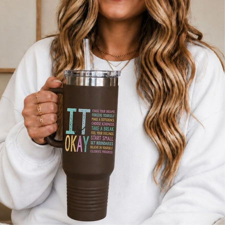 A woman in a white sweatshirt holds a dark brown insulated tumbler with a clear lid and straw. The tumbler has colorful text that reads 'IT OKAY' with smaller motivational text underneath.