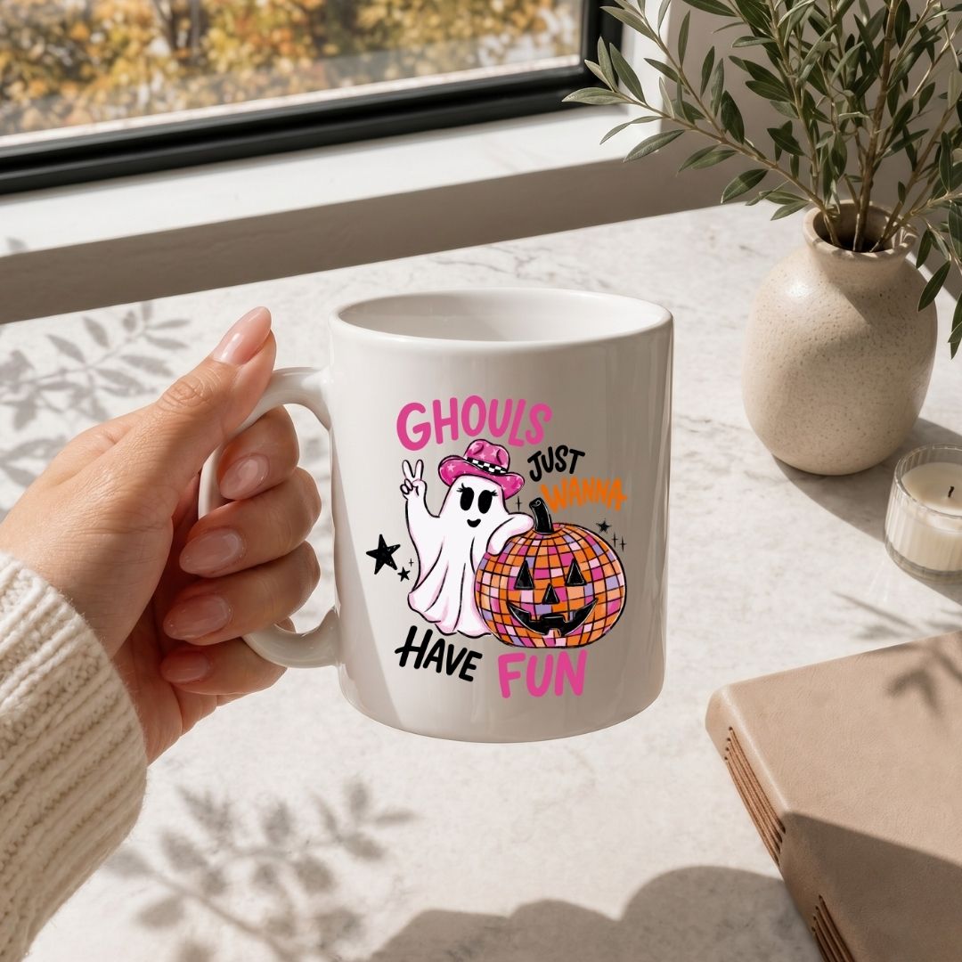 A person holding a white ceramic coffee mug printed with the Ghouls Just Wanna Have Fun Halloween illustration, set on a marble countertop next to a vase of greenery.