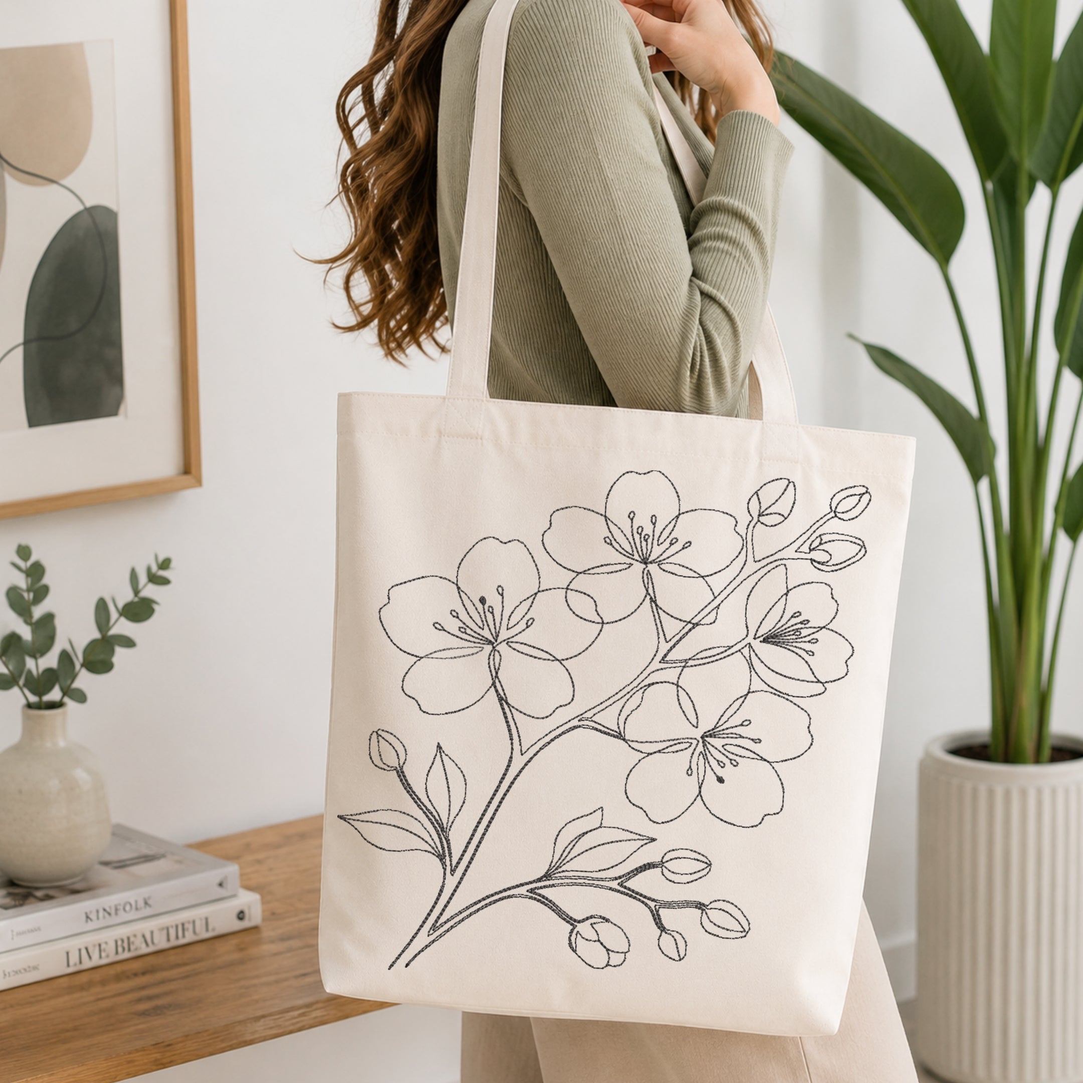 A woman wearing a sage green ribbed sweater carries a cream canvas tote bag with black line art of cherry blossoms. The scene is set in a bright, modern room with a wooden table, a potted plant, and a framed print.