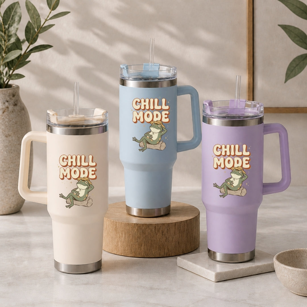 Three insulated travel tumblers with handles and straws are displayed on a tabletop in a styled indoor setting. The tumblers are in cream, light blue, and lavender, each printed with the words “CHILL MODE” and a frog illustration.