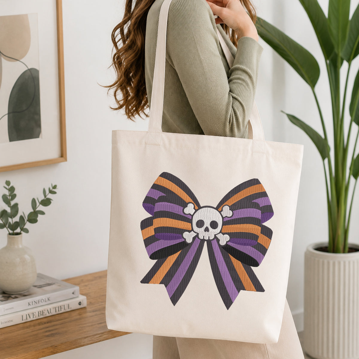A person wearing a green long-sleeve top and carrying a natural canvas tote bag printed with an embroidered purple, orange, and black striped bow featuring a skull and crossbones center.