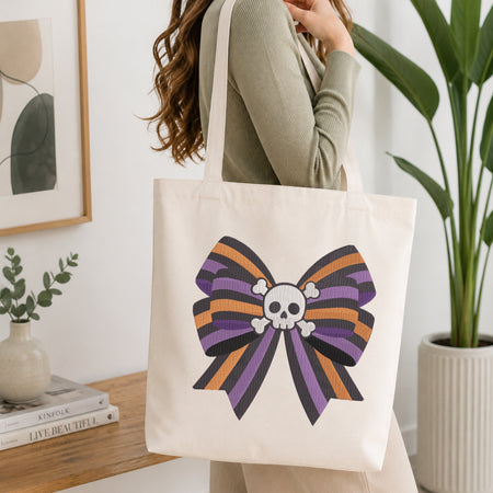 A person wearing a green long-sleeve top and carrying a natural canvas tote bag printed with an embroidered purple, orange, and black striped bow featuring a skull and crossbones center.