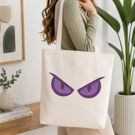 A woman wearing a sage green ribbed long-sleeve top carries a cream canvas tote bag featuring a graphic of two angry purple eyes. The setting is a bright, modern room with a wooden table, framed art, and a potted green plant.