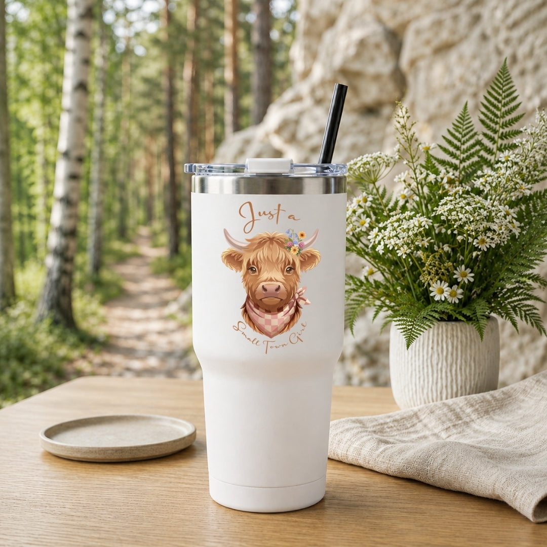 A white insulated tumbler with a clear lid and black straw stands on a wooden table. The tumbler is printed with a cute brown highland cow illustration and the words 'Just a Scottish Farm Cat.