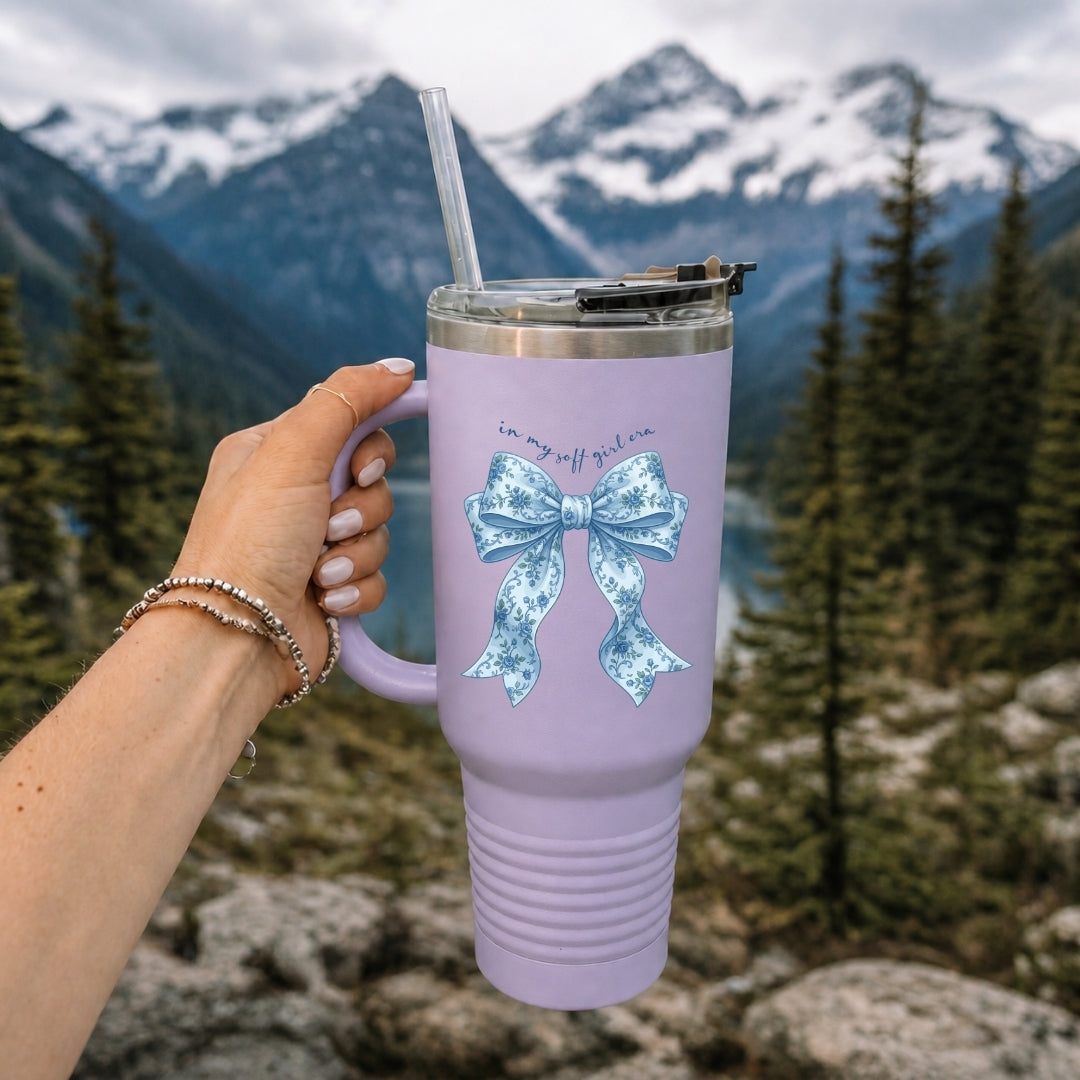 A person holding a lavender stainless steel tumbler with a straw, decorated with a blue floral bow graphic and the text "in my soft girl era."