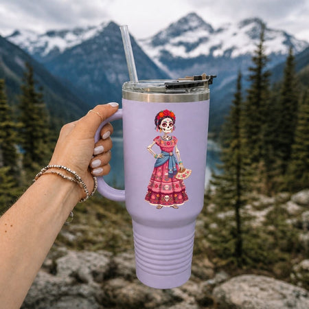 A hand holds a lavender insulated tumbler with a clear straw in front of a mountain and forest background. The tumbler features a colorful illustration of a Day of the Dead (Dia de los Muertos) woman in a pink and red dress.