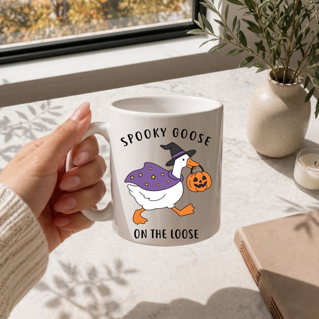 A person holding a white ceramic coffee mug printed with the Spooky Goose on the Loose Halloween illustration, placed on a marble surface next to a vase of greenery.