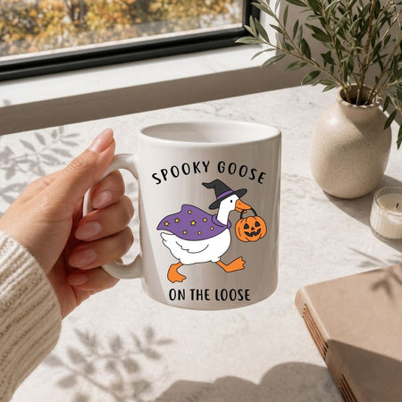 A person holding a white ceramic coffee mug printed with the Spooky Goose on the Loose Halloween illustration, placed on a marble surface next to a vase of greenery.