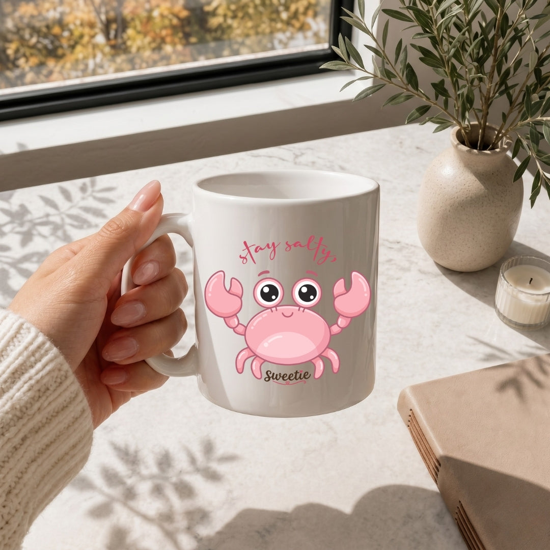 Person holding a mug with a pink crab design and text, sitting at a table with a window and plant in the background.