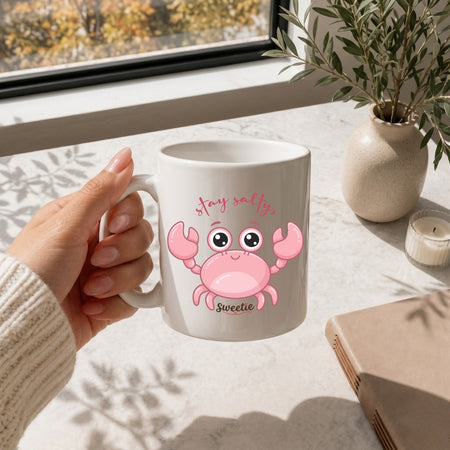 Person holding a mug with a pink crab design and text, sitting at a table with a window and plant in the background.