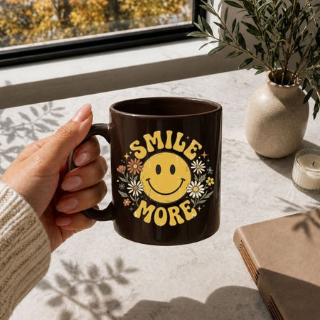 A close up of a hand holding a glossy brown ceramic mug printed with a vintage yellow smiley face graphic and floral accents.