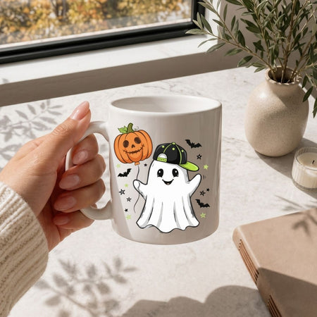 A hand holding a white ceramic mug decorated with a cute cartoon ghost wearing a green and black cap, next to a carved orange pumpkin, with a window and potted plant in the background.