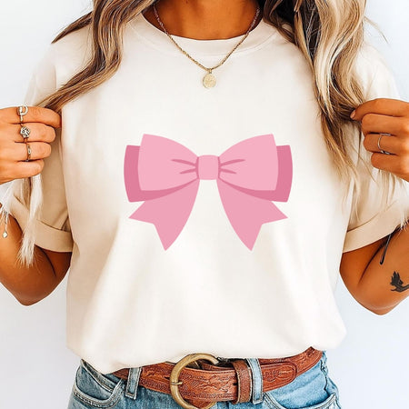 A woman wearing a cream-colored short-sleeve t-shirt with a large pink bow graphic printed on the chest, paired with blue jeans and a brown leather belt.