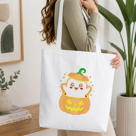 Woman carrying a white natural canvas tote bag featuring an embroidered ghost inside a jack-o-lantern pumpkin design in a bright, modern interior setting