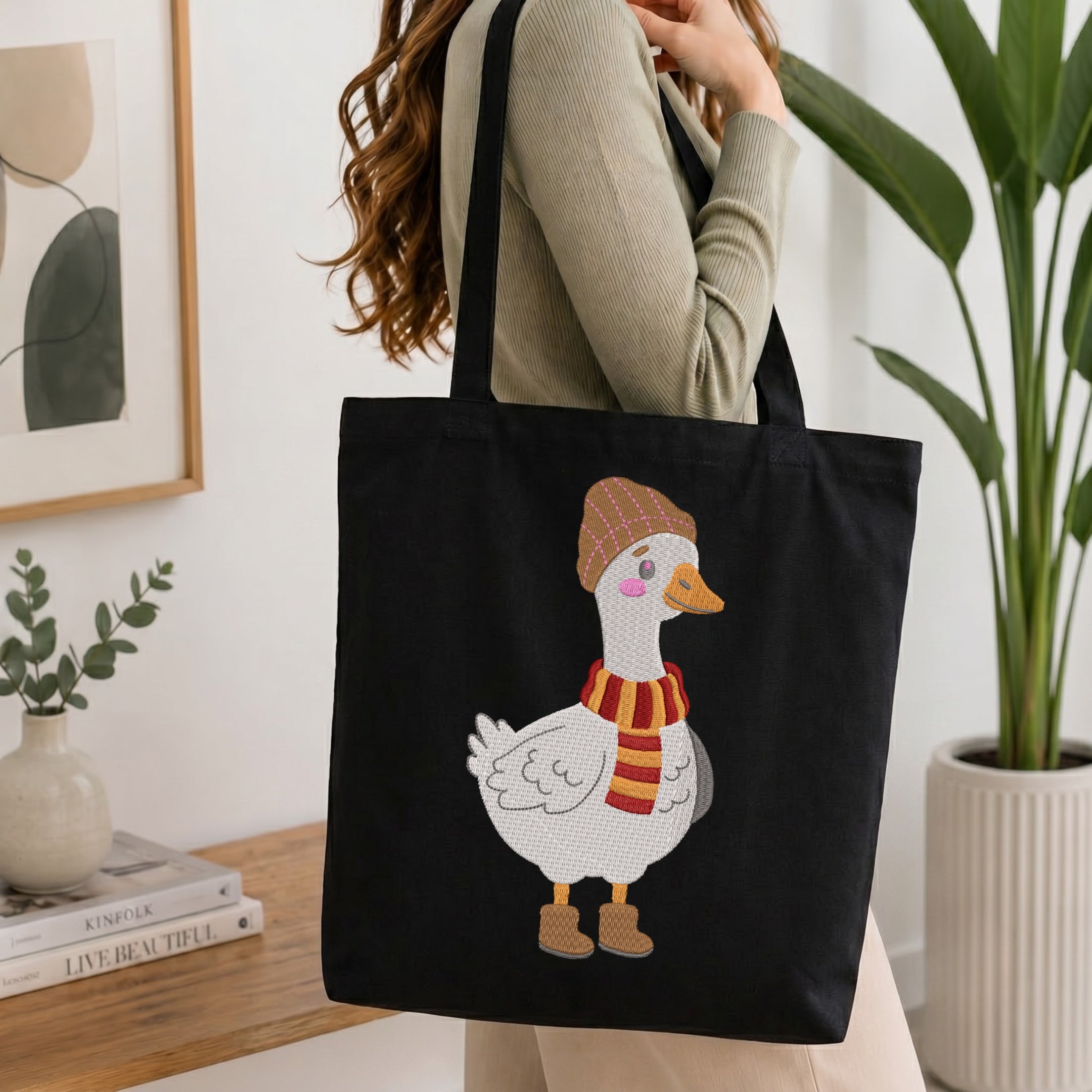 A person wearing a beige ribbed sweater carries a black canvas tote bag with a white goose illustration wearing a brown hat and red-and-yellow striped scarf, shown in a bright indoor room with plants and framed art.