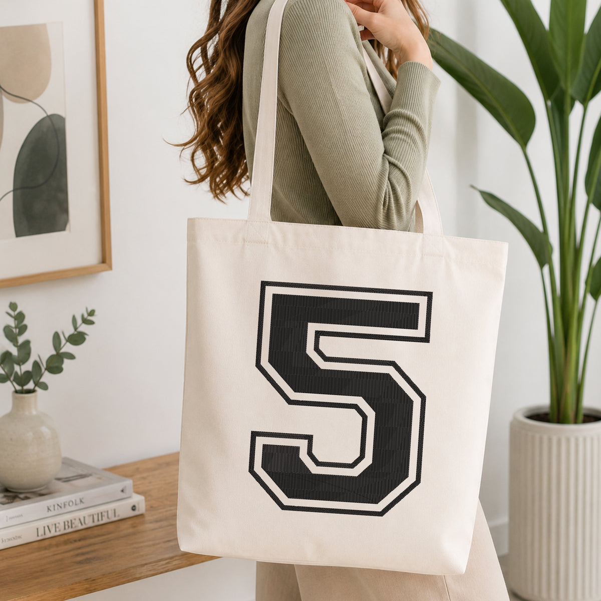 Varsity number "5" machine embroidery design in black with white outline stitched on a natural canvas tote bag held by a person in a modern interior setting.