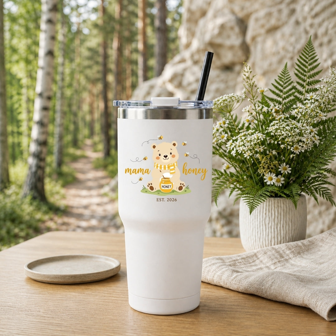 A white insulated tumbler with a clear lid and black straw stands on a wooden table in a forest setting. The tumbler is printed with a cute bear illustration, the words 'mama honey', and 'EST.