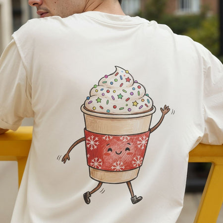 A man wearing a cream-colored t-shirt, viewed from behind. The back of the shirt features a large, hand-drawn style graphic of a cute, smiling whipped coffee drink character with arms, legs, and colorful sprinkles.