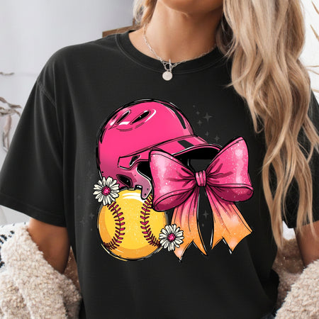 A woman wearing a black graphic t-shirt featuring a pink softball helmet, a large pink ribbon bow, a yellow softball, and white daisies.